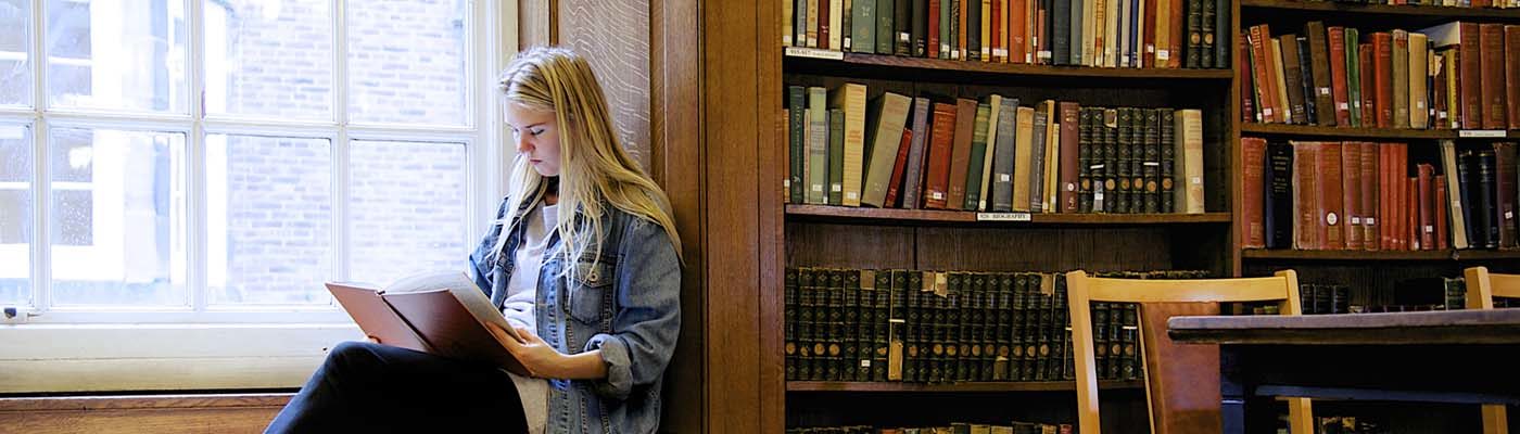 A student sits on a window seat reading a book in a library.