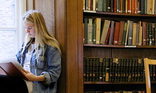 A student sits on a window seat reading a book in a library.