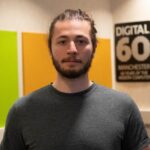 Person in a grey t‑shirt standing in a modern indoor space with colourful wall panels and a ‘Digital 60 Manchester’ sign in the background