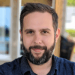 A person with short dark hair and a beard is wearing a dark button-up shirt while standing in front of a wooden structure with blurred outdoor scenery behind them.
