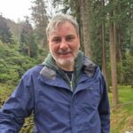 Person standing outdoors wearing a blue jacket, with tall evergreen trees and greenery in the background