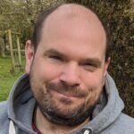 A person wearing a grey hoodie is standing outdoors beside a large tree trunk, with green grass and several small trees visible in the background.