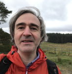 A person wearing an orange outdoor jacket stands in a grassy rural landscape with rolling hills, trees, and a cloudy sky in the background