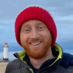 A person wearing a red knit hat and a dark puffer jacket is standing on a sandy beach, with a lighthouse and cloudy blue sky in the distance.