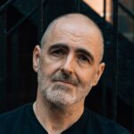 Person standing outdoors in front of a metal staircase, wearing a black t‑shirt and jeans with arms crossed and sunglasses hanging from the shirt.