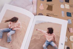 A photograph of an open book with the same model in a pink top and jeans on the beach in each picture. Behind there is a board with illustrations of garments next to correlating fabric swatches and their material composition.