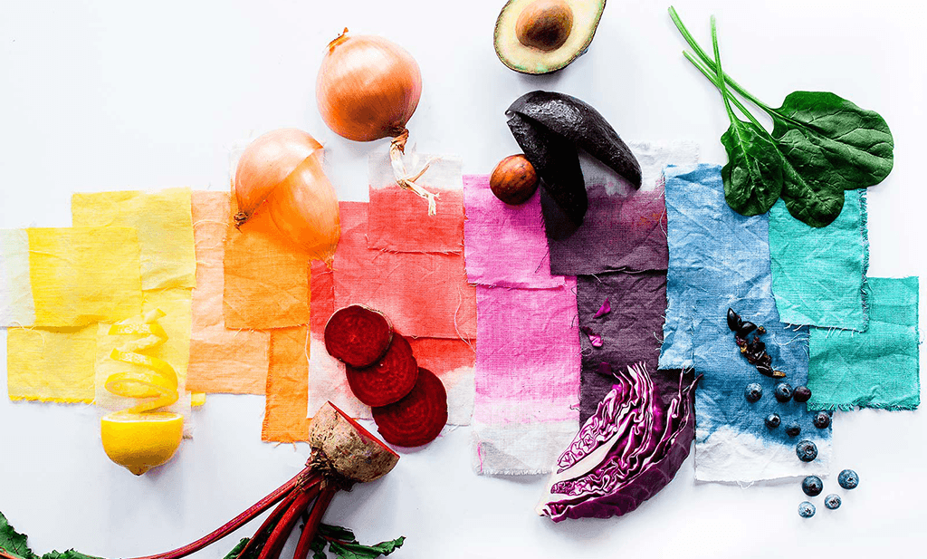 An image with lots of fabric displayed that has been dyed naturally by various vegetables that are also shown in the photo (beetroot, onion etc).