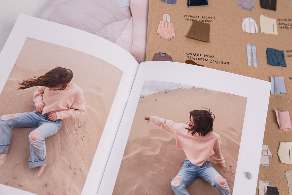 A photograph of an open book with the same model in a pink top and jeans on the beach in each picture. Behind there is a board with illustrations of garments next to correlating fabric swatches and their material composition.