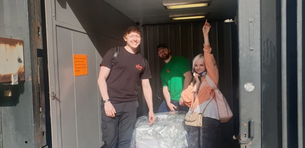 Two male presenting people and a female presenting person stand next to a very large package covered in foil  in a grey boxed space. 