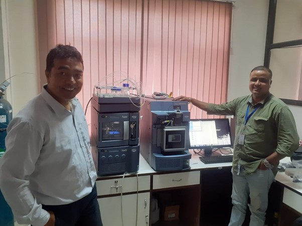 Two male presenting people standing next to two pieces of eletronic equipment (mass spectrometer equipment) in front of a shuttered window.