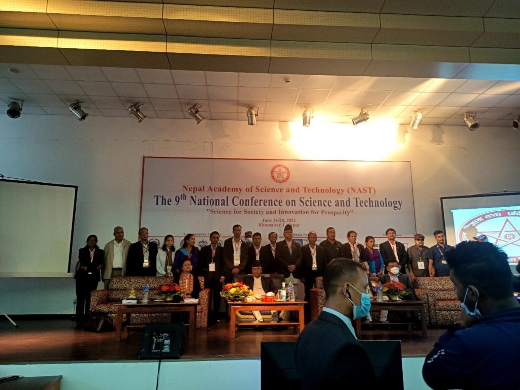 A group of people are standing on a platform in a conference setting behind a panel of seated people. The banner at the back reads 'the 9th Nationa conference on Science and Technology'