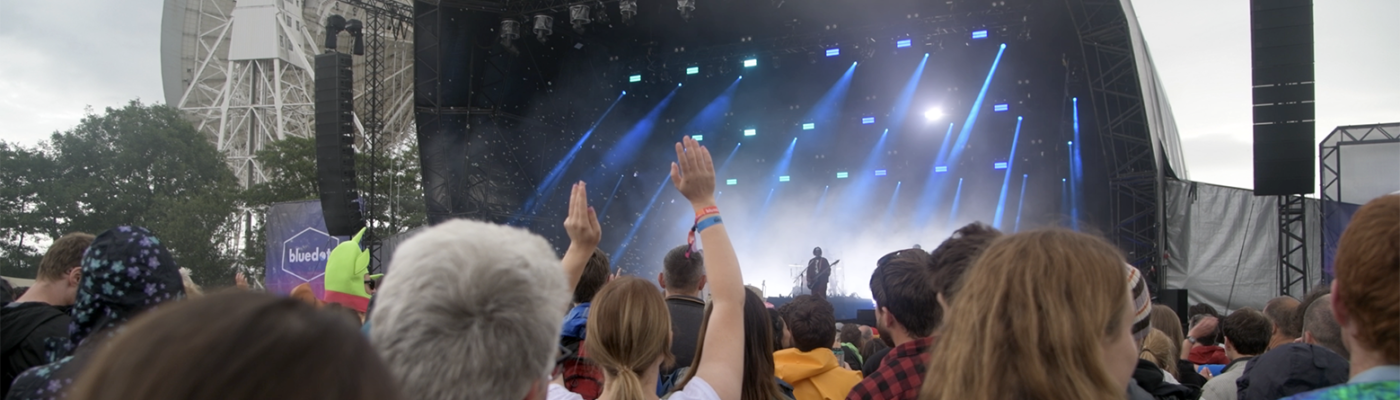 A photo of the main stage of bluedot. The busy crowd is in the foreground with hands raised and people dancing. On stage is a light show with a band's frontman singing.