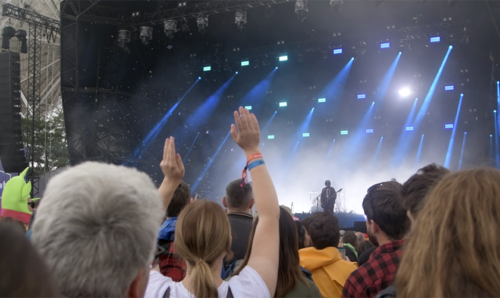 A photo of the main stage of bluedot. The busy crowd is in the foreground with hands raised and people dancing. On stage is a light show with a band's frontman singing.