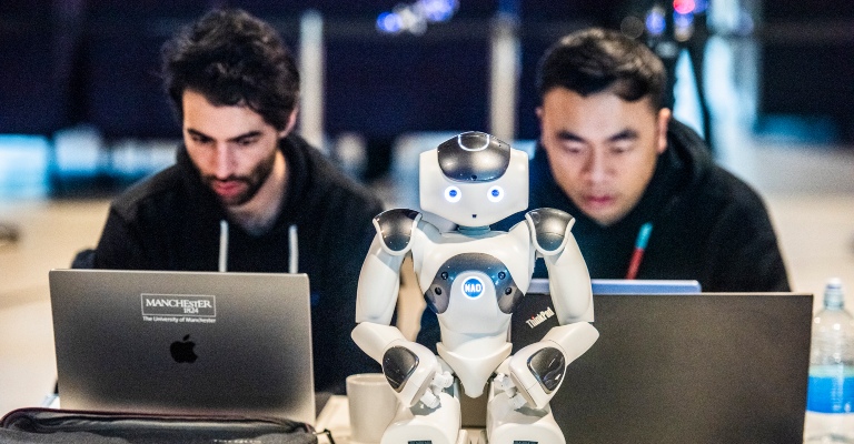 A small robot in the foreground looks at the camera, while two researchers in the background look intently at their laptops.