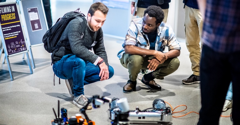 Two men crouch while watching two moving robots on the floor.