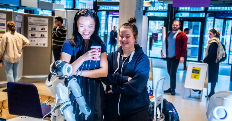 Two women smile while looking at a small robot on a desk.