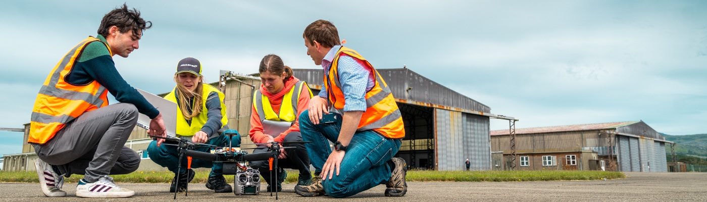Four students in high-vis crouch down to inspect their drone.