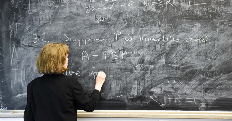 A teacher writes on a blackboard with her back to the camera.