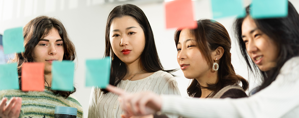 Four students looking at post-it notes on a wall.