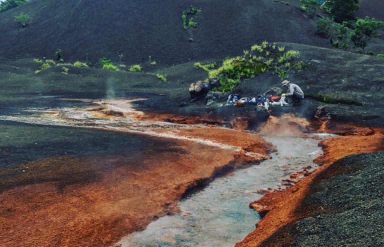 Person squatting next to hot springs on a volcano.